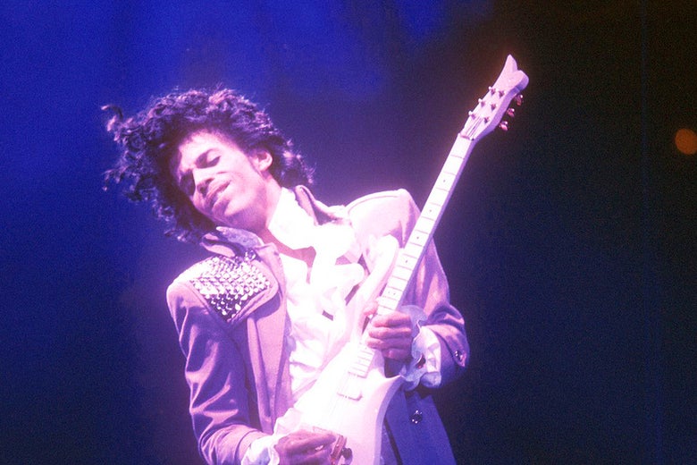 Prince, bathed in purple light, holds a guitar.